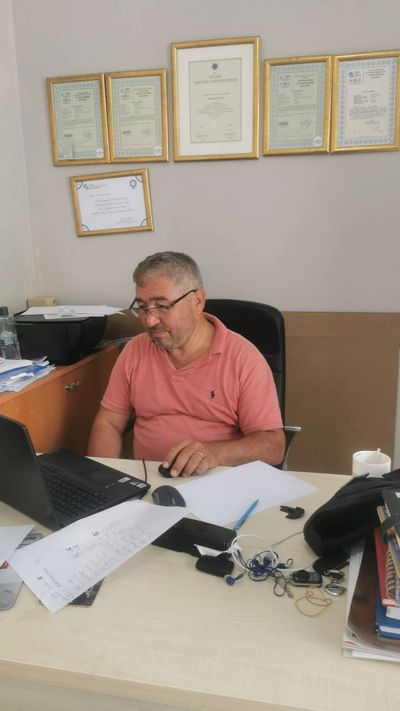 Man working on a laptop at a cluttered office desk with certificates on the wall.