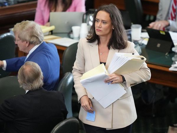 A woman in a beige blazer holding papers in a busy office setting.