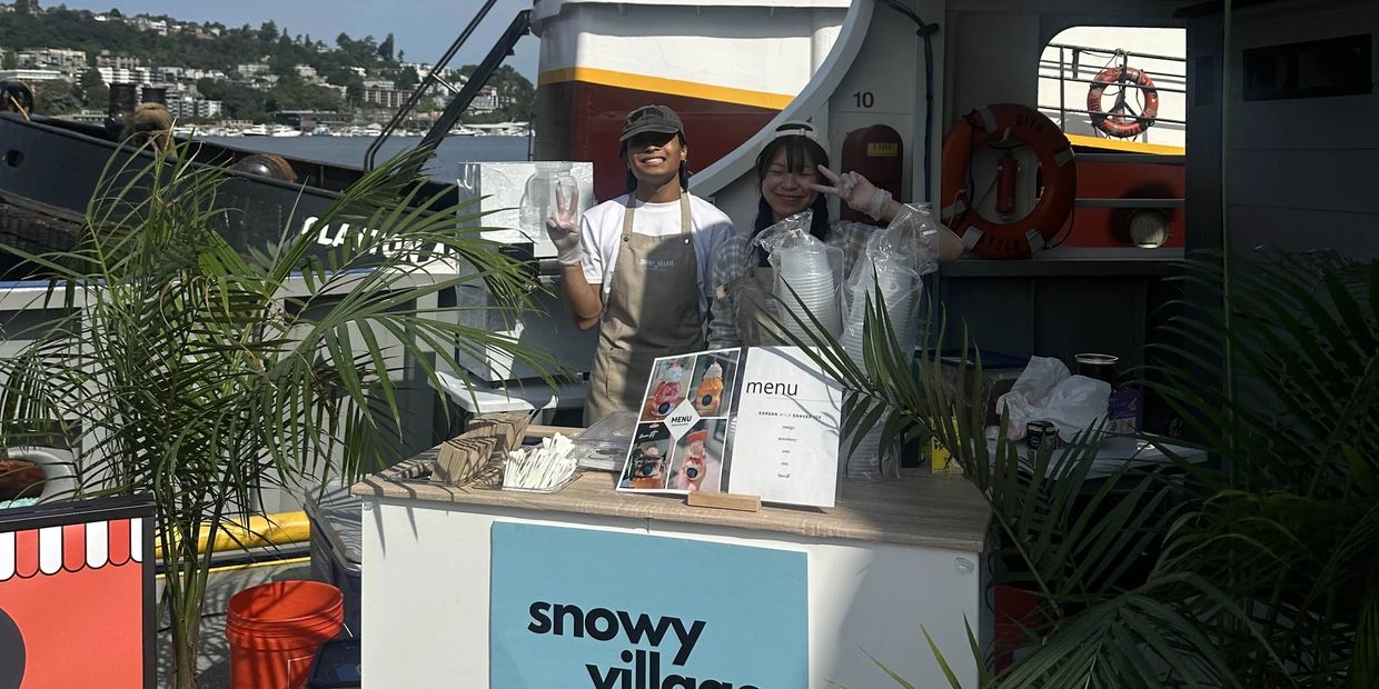 Two smiling vendors at a snowy village shaved ice stand on a boat deck.