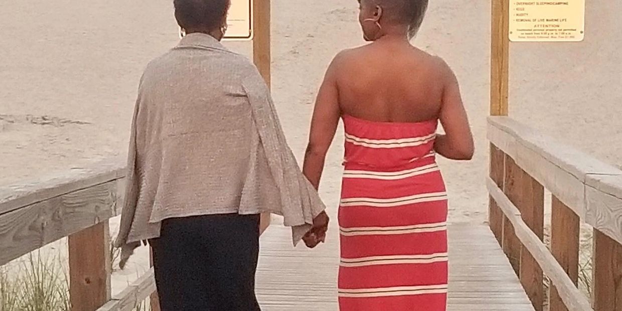 Two women holding hands on a boardwalk leading to the beach at sunset.