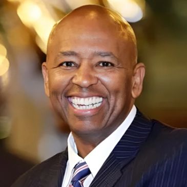 A smiling man in a suit and tie with a bright background.