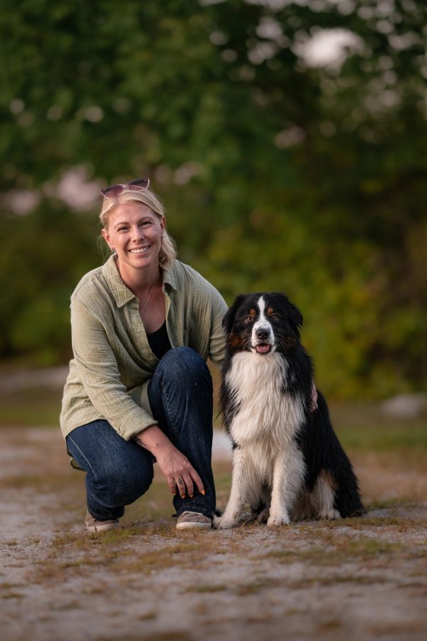 A smiling woman crouches beside a happy black and white dog outdoors.