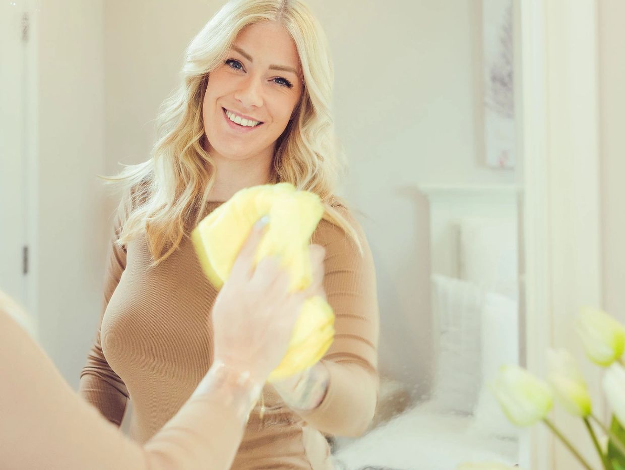 Smiling woman cleaning a mirror with a yellow cloth.