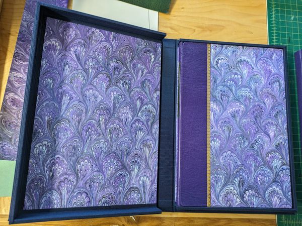 An open decorative box with a purple marbled-patterned book inside on a wooden surface.