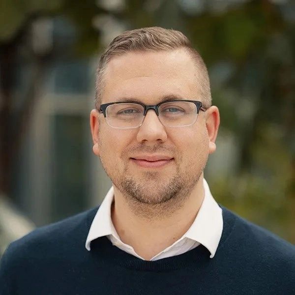 Portrait of a man with glasses smiling softly outdoors.