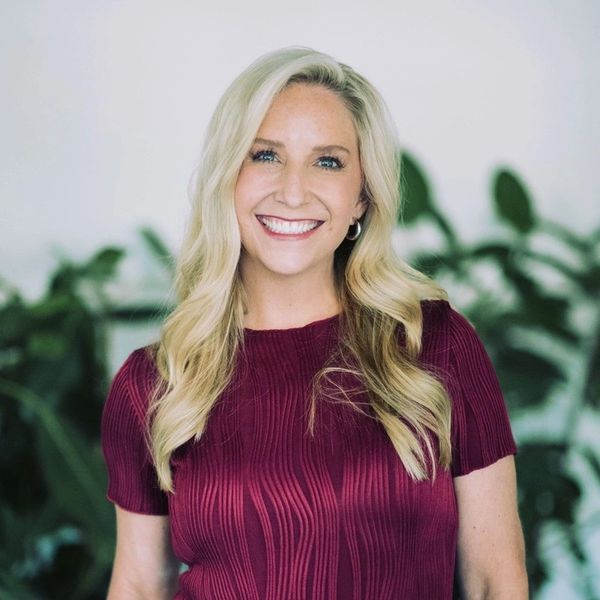 Smiling woman with long blonde hair wearing a maroon top, standing in front of green plants.