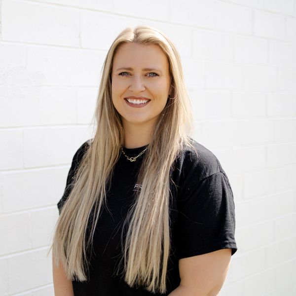 Smiling woman with long blonde hair in a black shirt against a white brick wall.