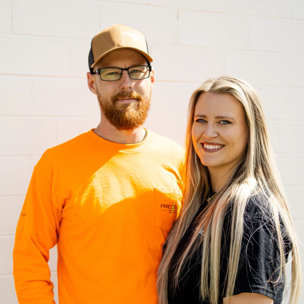A man and woman smiling together outdoors against a white wall.
