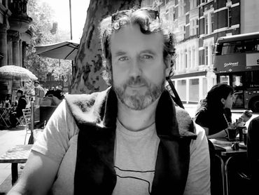 Man with curly hair and beard sitting outdoors at a café.