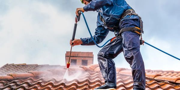 Worker pressure washes roof tiles wearing safety gear.