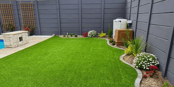 A backyard with artificial grass, flower beds, and a gray fence.