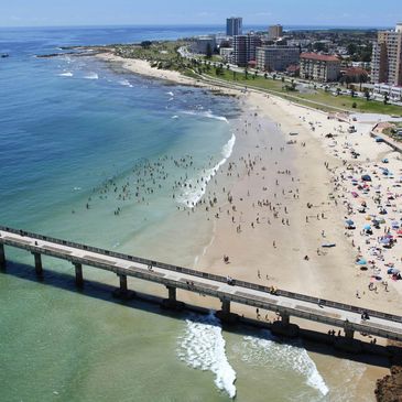 A crowded beach with a pier and buildings in the background.