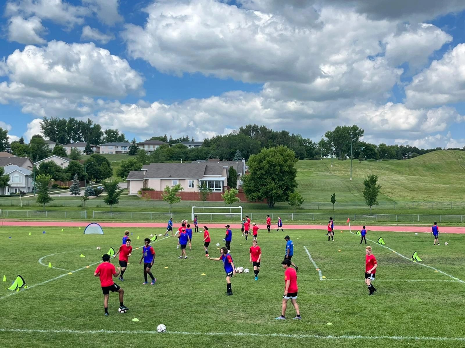 Youth soccer players in red and blue practicing on a green field with suburban houses and cloudy sky.