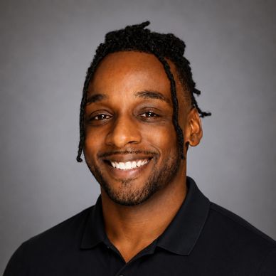Smiling man with braided hair wearing a black polo shirt against a gray background.