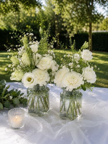 Two jars with white flowers and a candle on a table outdoors.