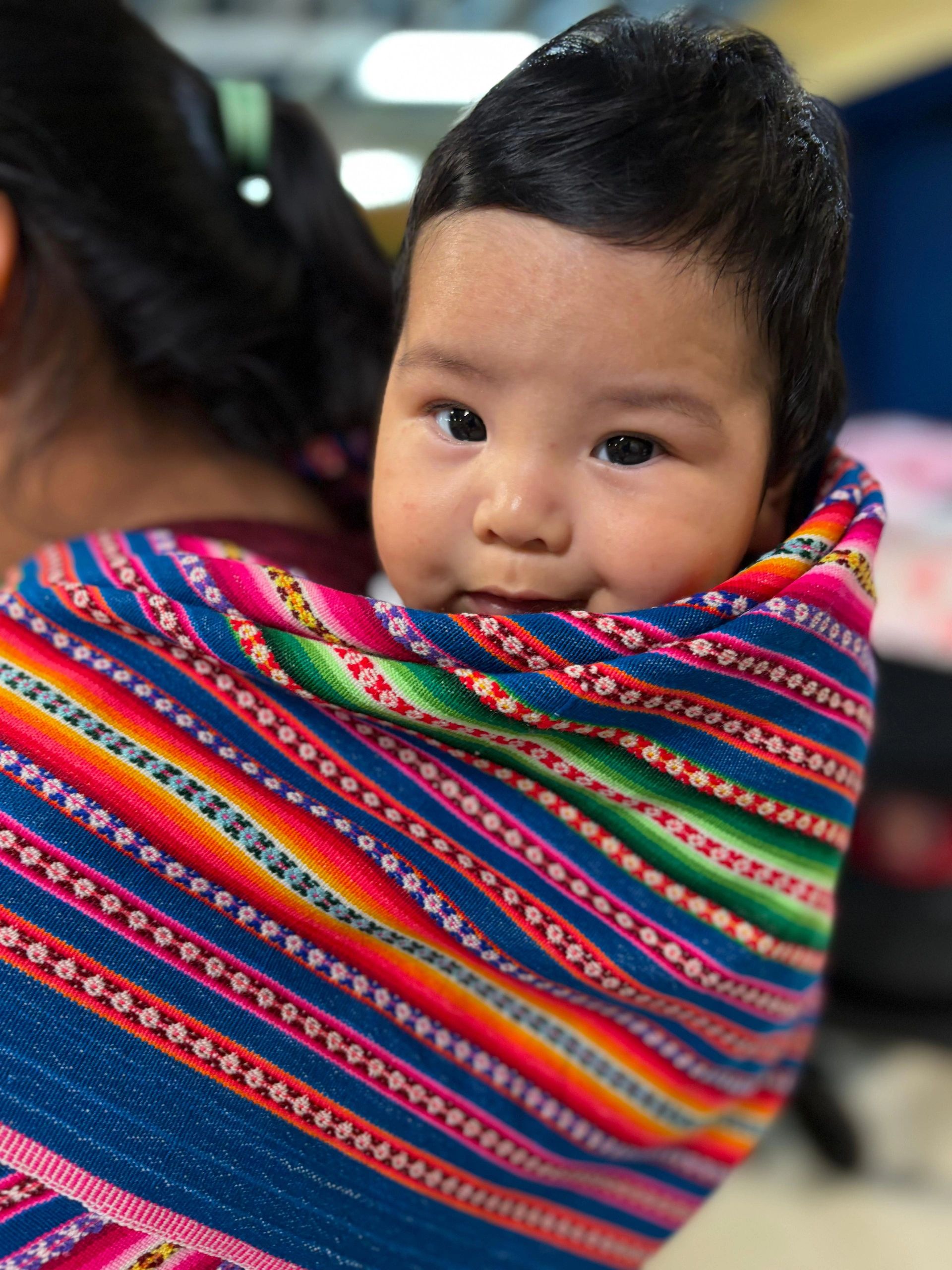 An image of a baby being carried on it's mother's back in a brightly colored carrier.