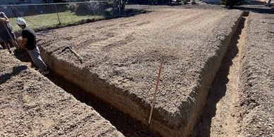 Two people working on a large dug-out trench in a residential yard under clear skies.