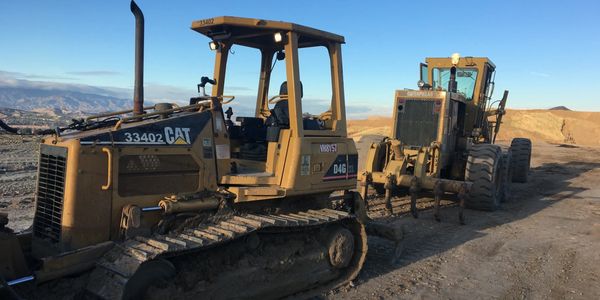 Two Caterpillar construction vehicles on a dirt road under a clear blue sky.