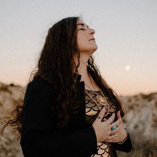 A woman with long curly brown hair and eyes closed raises her head to the sky as the sun sets with t