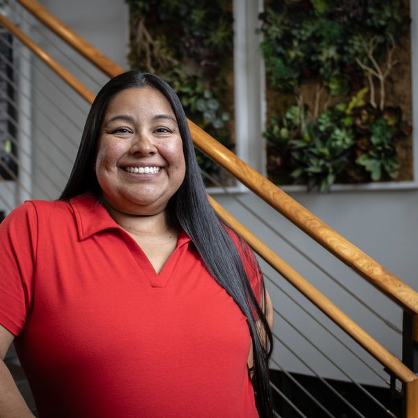 Smiling woman in a red shirt standing by a staircase with greenery on the wall.