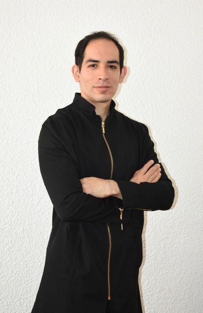Man in black jacket with crossed arms stands against white wall.