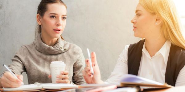 Two women discussing work with notebooks and coffee.