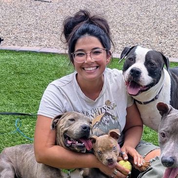 A smiling woman surrounded by happy dogs outdoors on grass.