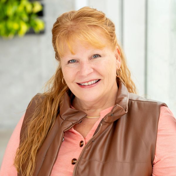 Smiling woman with red hair wearing a brown vest and pink shirt.