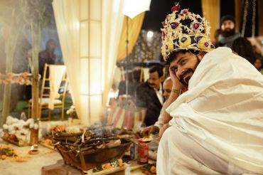 A man in a ceremonial crown and white robe participates in a ritual with incense and offerings.