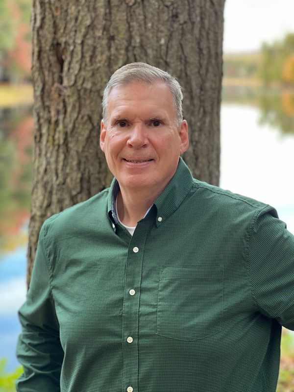 Man in a green shirt standing by a tree near a lake.