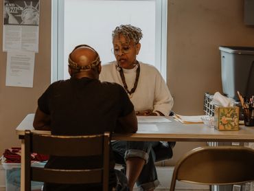 Two people engaged in a serious conversation at a table in a modest room.