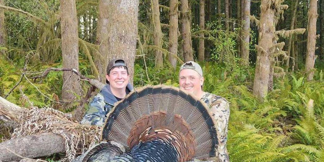 Two men in camouflage hold a large turkey in a forest.
