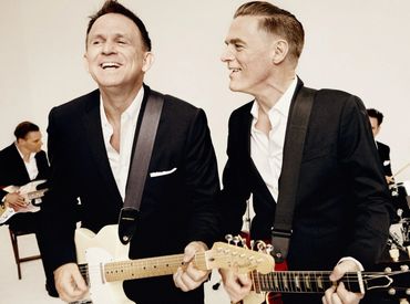 Two men in suits playing electric guitars and smiling.