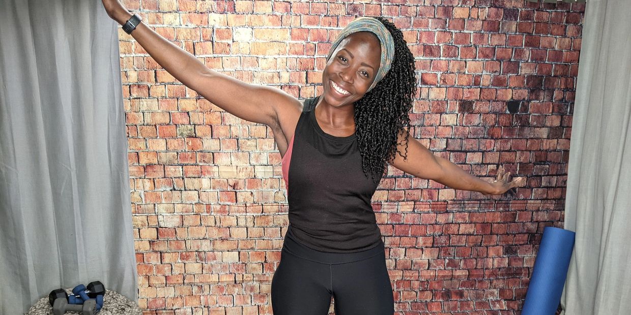 Smiling woman in workout attire stretching arms in front of a brick wall.