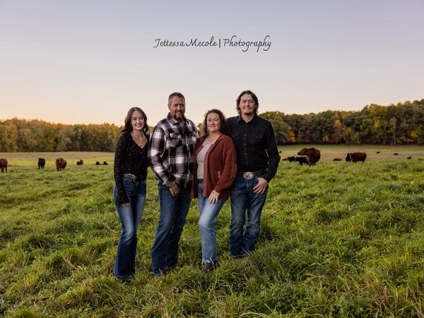 Four people posing in a grassy field with cattle in the background.
