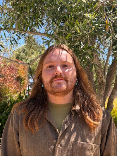 Man with long hair standing outdoors near a tree.