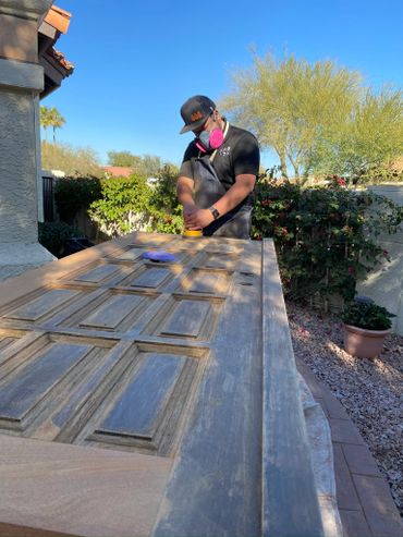 Person sanding a wooden door outdoors on a sunny day.