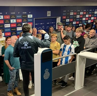 A group of fans listens to a Kelme representative at a sports venue.