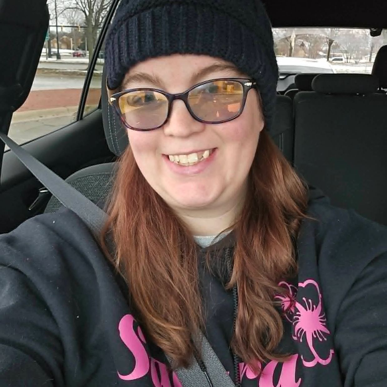 Smiling woman wearing glasses and a beanie inside a car with a seatbelt on.