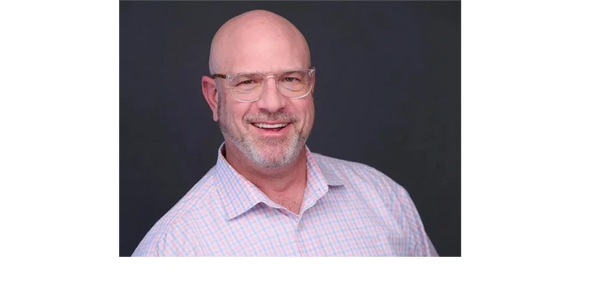 Smiling bald man with glasses wearing a checkered shirt against a dark background.
