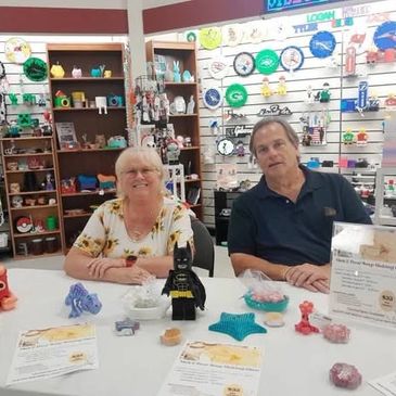 Two people sitting at a table displaying various handmade crafts and a Batman figure.