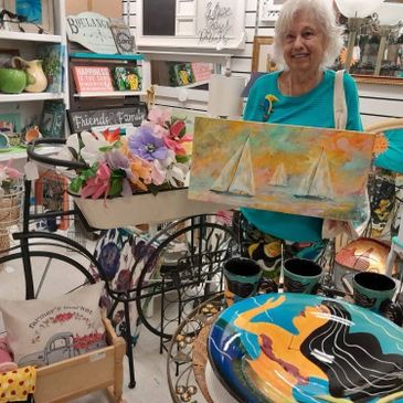 Elderly woman holding a sailboat painting in a colorful gift shop.