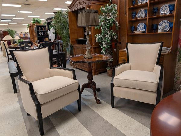 Two beige armchairs with dark wooden frames flank a small ornate table with a lamp in a furniture store.