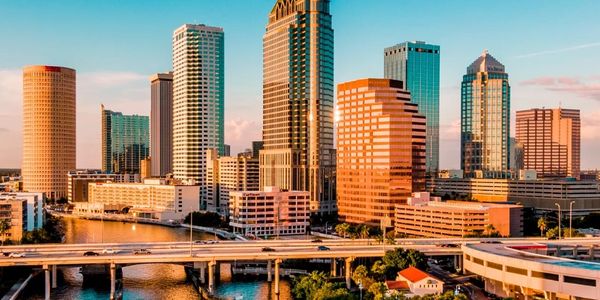 Sunlit downtown Tampa, FL skyline with buildings and a river bridge.