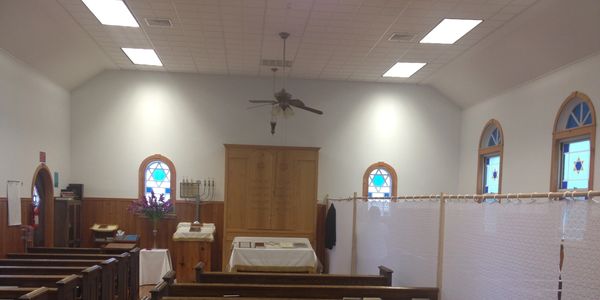 Interior of a small synagogue with wooden pews and stained glass windows.