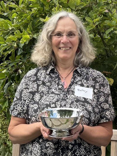 Smiling woman named Betsy holding a shiny silver bowl outdoors.