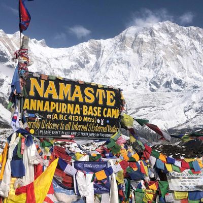 Colorful prayer flags and a sign welcome visitors to Annapurna Base Camp with snowy mountains in the background.
