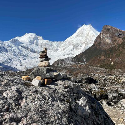 Stacked stones on rocky terrain with snowy mountains and clear blue sky.