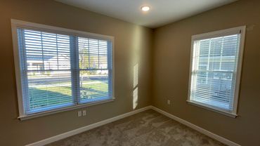 Empty room with beige walls and carpet, two windows with blinds letting in natural light.