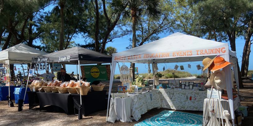 Outdoor market stalls with crafts and baked goods under trees.